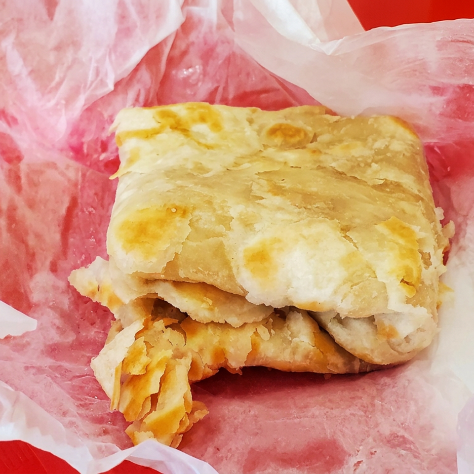 Golden flaky stuffed pastry wrapped in pink paper, with a small container of red dipping sauce beside it.